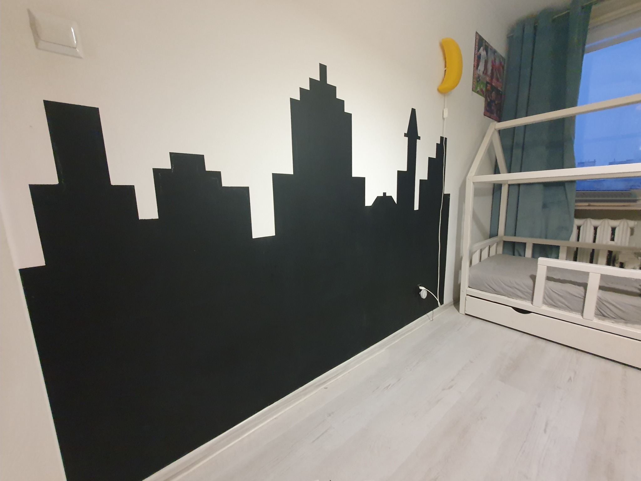 Czarny, ręcznie malowany motyw panoramy miasta na białej ścianie w pokoju dziecięcym, obok łóżka w kształcie domku z szarej pościeli i lampki nocnej w kształcie banana.