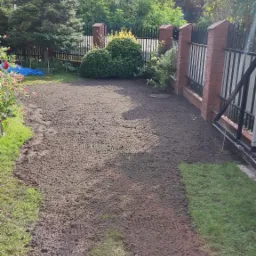 Przygotowany teren pod trawnik z widocznymi śladami po bronowaniu, obrzeża z trawy i ogrodzenie z ceglanymi słupkami w tle.