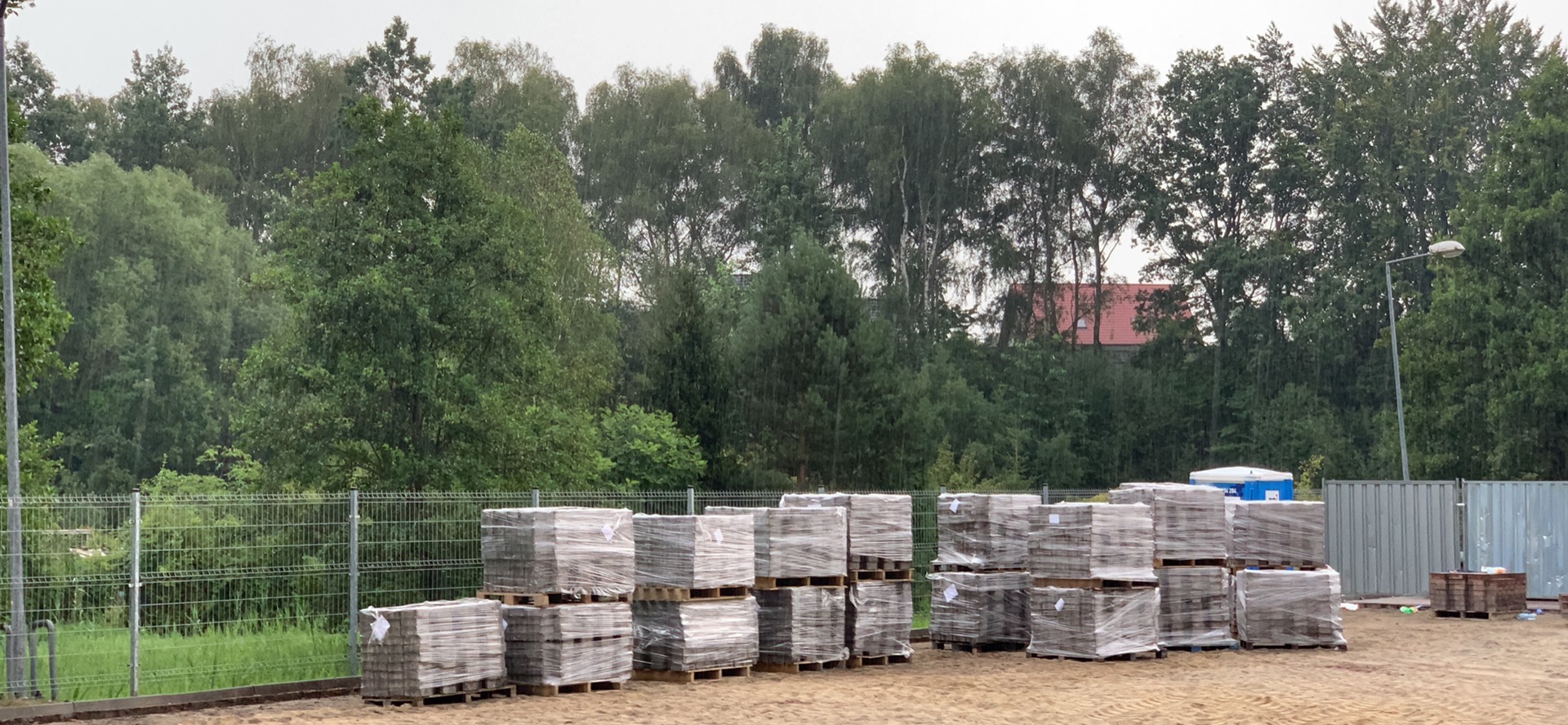 Kostka brukowa ułożona na paletach, zabezpieczona folią, składowana na placu budowy, w tle zieleń drzew i fragment budynku z czerwonym dachem.