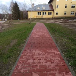 Czerwony chodnik z kostki brukowej w kształcie litery H prowadzi w kierunku żółtego budynku z szarym dachem, otoczony trawnikiem i drzewami w pochmurny dzień.