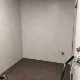 Wnętrze łazienki w trakcie prac wykończeniowych: beżowe płytki ścienne, ciemnobrązowa podłoga, widoczne puszki elektryczne i fragment odkurzacza przemysłowego.