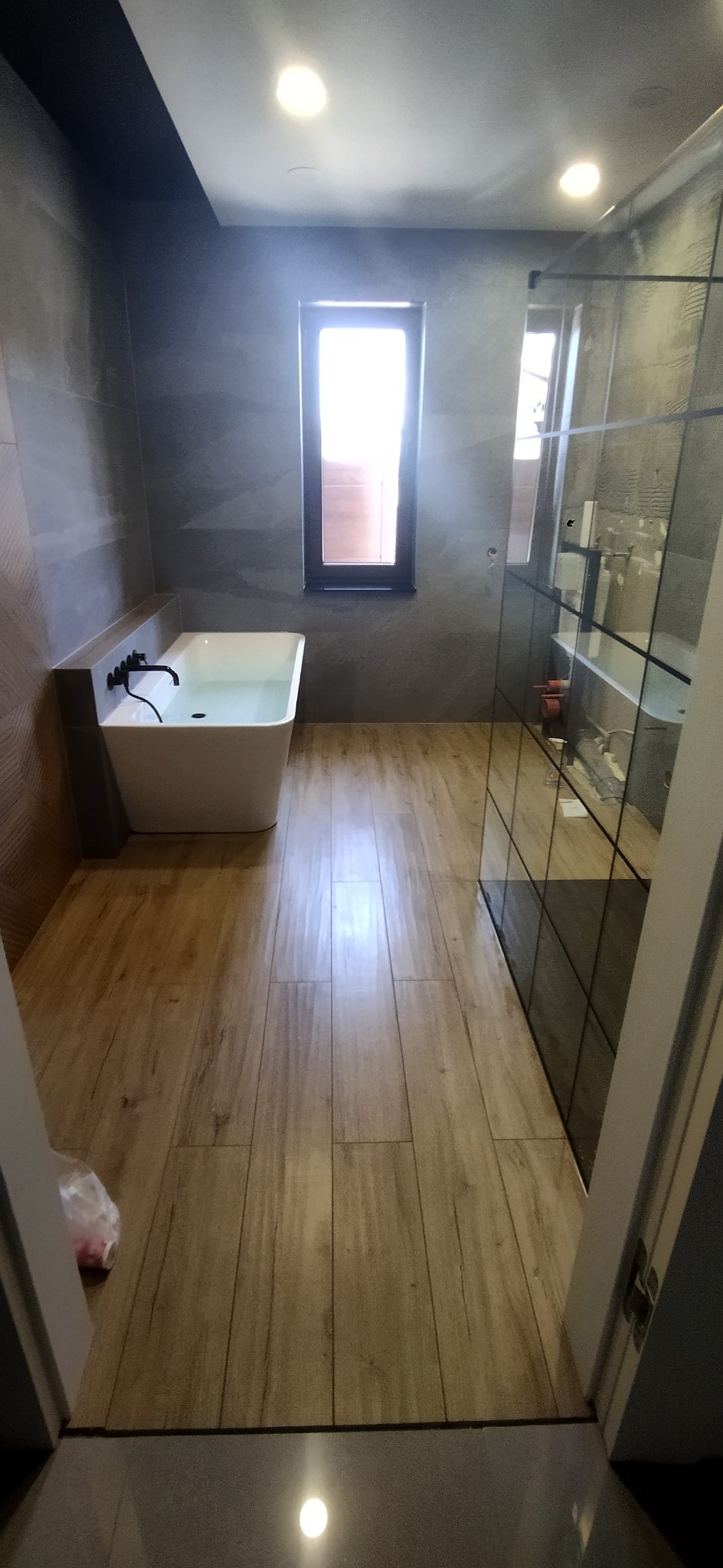 Nowoczesna łazienka z drewnopodobną podłogą, szarą ścianą z kamienia, białą wanną wypełnioną wodą i kabiną prysznicową ze szklanymi ściankami w czarnych ramach.