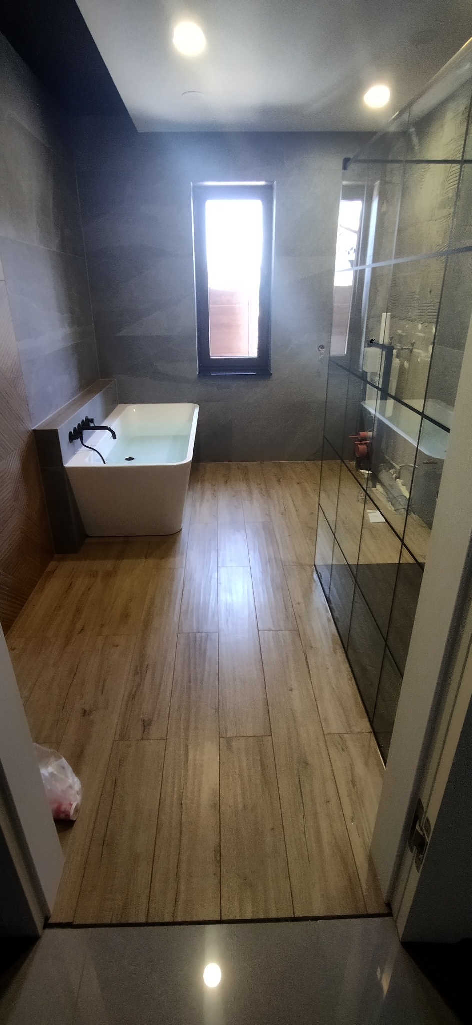 Nowoczesna łazienka z wolnostojącą wanną wypełnioną wodą, kabiną prysznicową typu walk-in z czarnymi profilami i drewnopodobną podłogą z płytek.