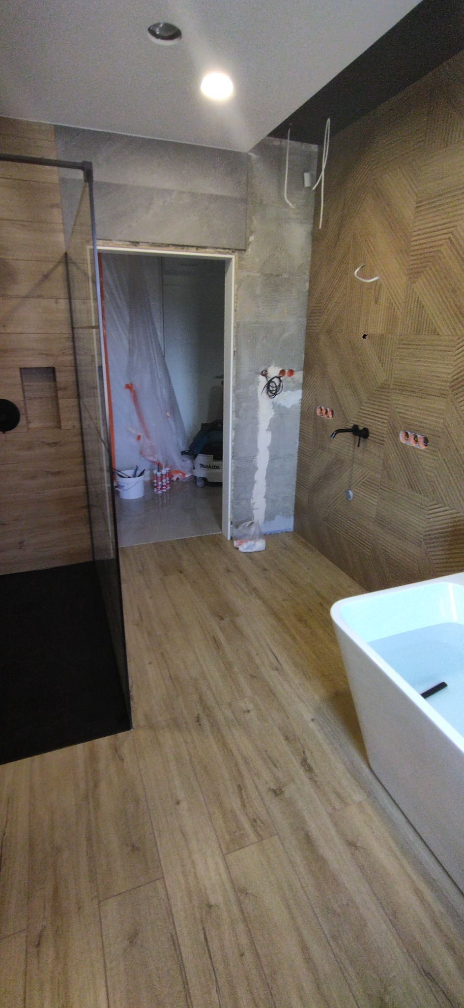 Łazienka w trakcie remontu: drewnopodobna podłoga, ściana z geometrycznym wzorem, częściowo wykończona kabina prysznicowa z czarnymi elementami, wolnostojąca wanna wypełniona wodą, widoczne puszki...