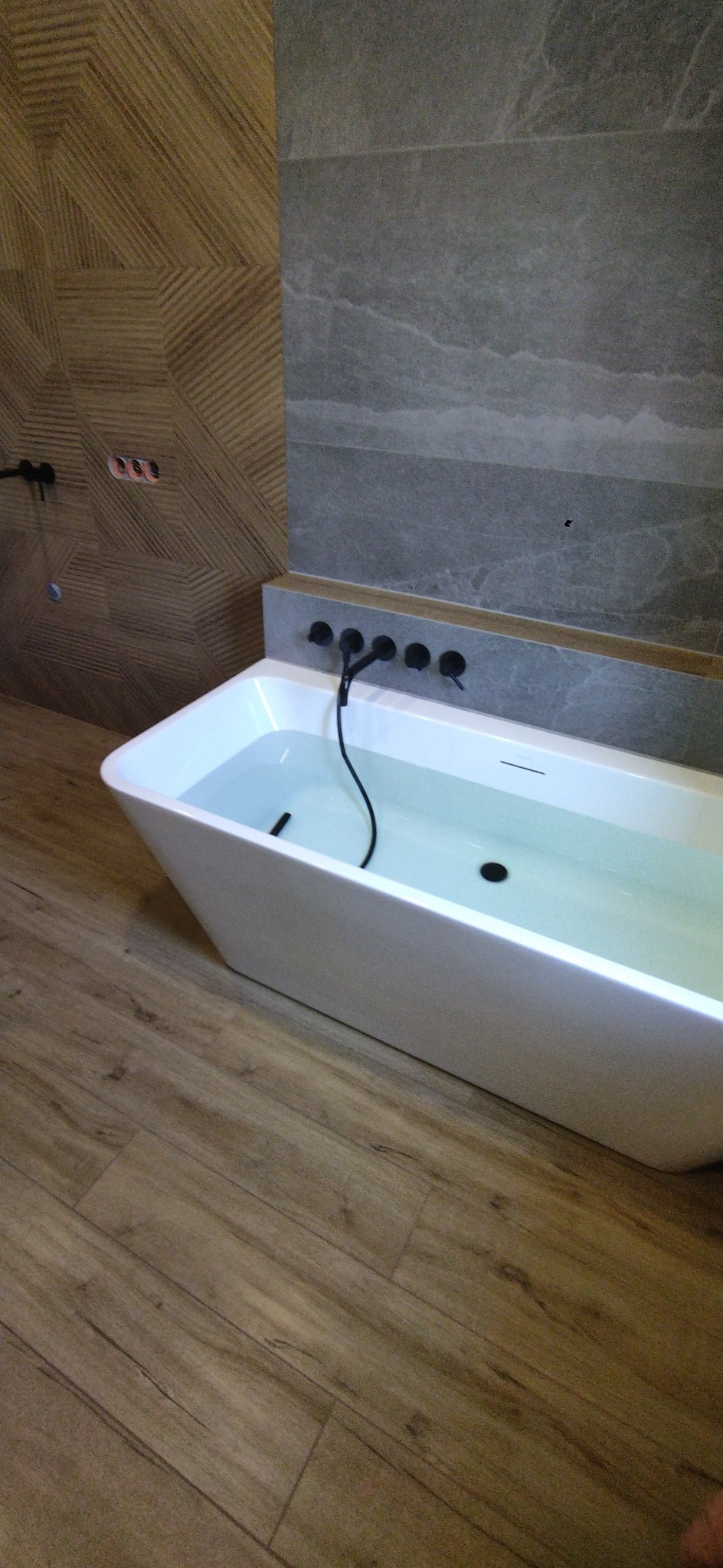 Nowoczesna wanna wypełniona wodą, z czarną armaturą w ścianie z szarych płytek imitujących kamień, obok ściana z dekoracyjnych paneli drewnopodobnych, na podłodze płytki imitujące drewno.