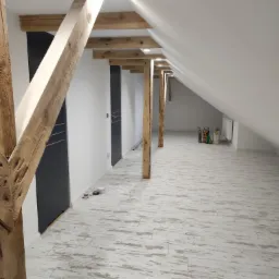 Wykończone poddasze z drewnianą konstrukcją stropu, białymi ścianami, szarymi drzwiami i podłogą z paneli imitujących postarzane drewno.