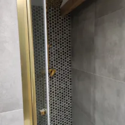 Nowoczesna kabina prysznicowa z złotymi detalami, heksagonalne mozaiki na ścianie i szara płytka wielkoformatowa, drewniany element stropu