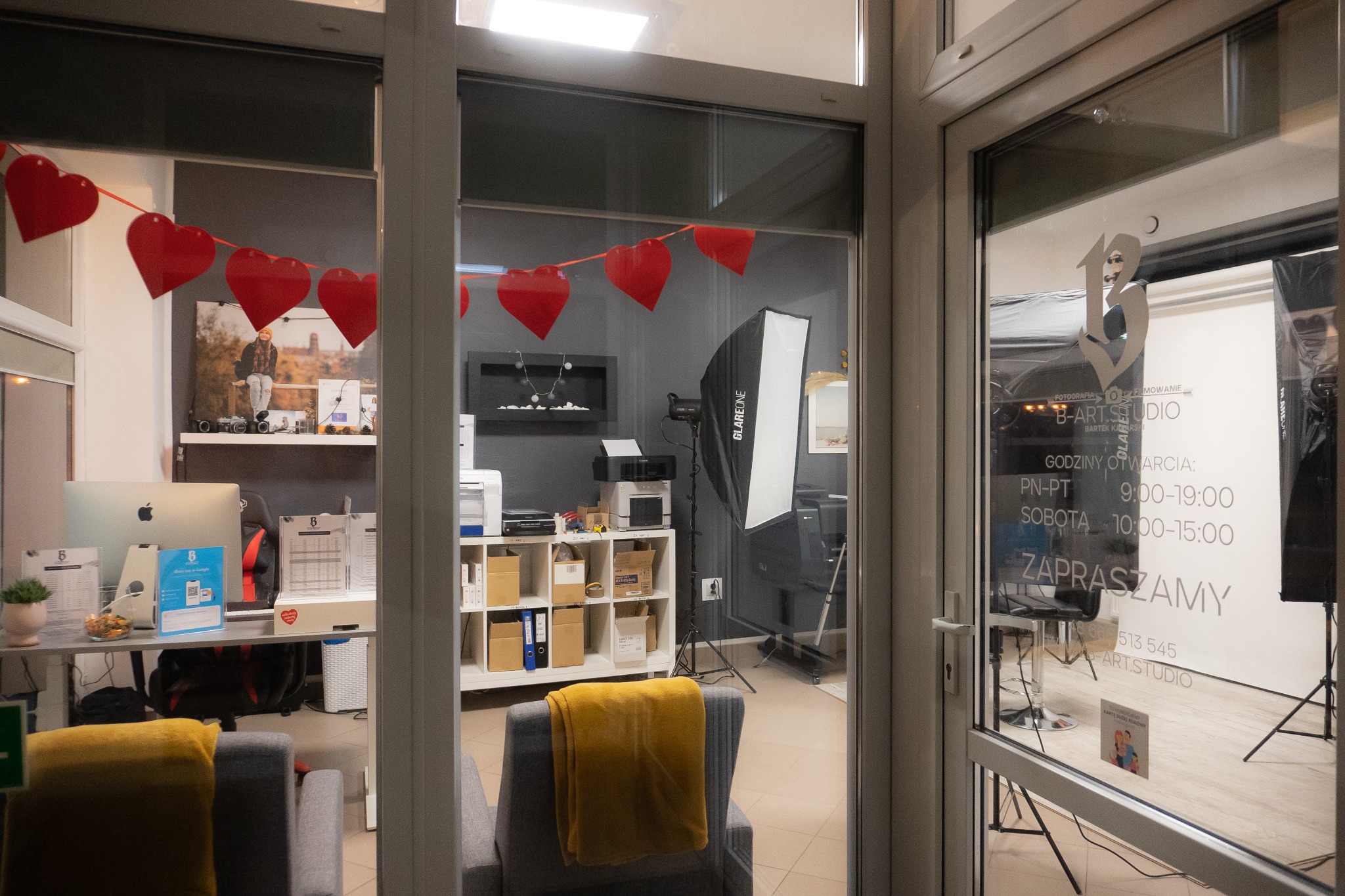 Wnętrze studia fotograficznego widoczne przez szybę, dekoracja z czerwonych serc, sprzęt oświetleniowy Glareone, biurko z komputerem Apple, godziny otwarcia na szybie.