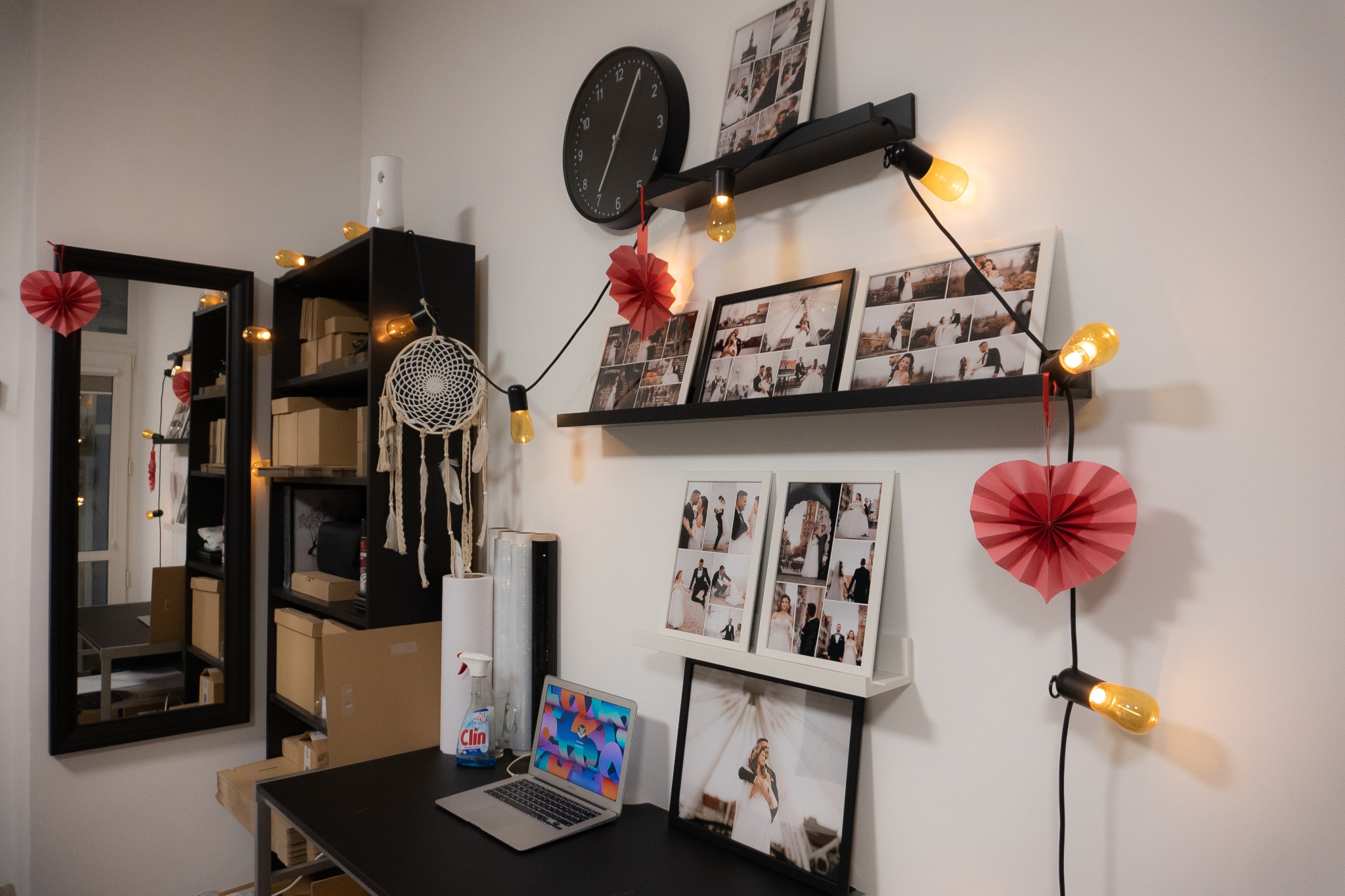 Ściana z dekoracjami: zdjęcia ślubne w ramkach na półkach, girlanda z żarówkami, zegar, lustro, łapacz snów, biurko z laptopem i płynem do czyszczenia Clin.