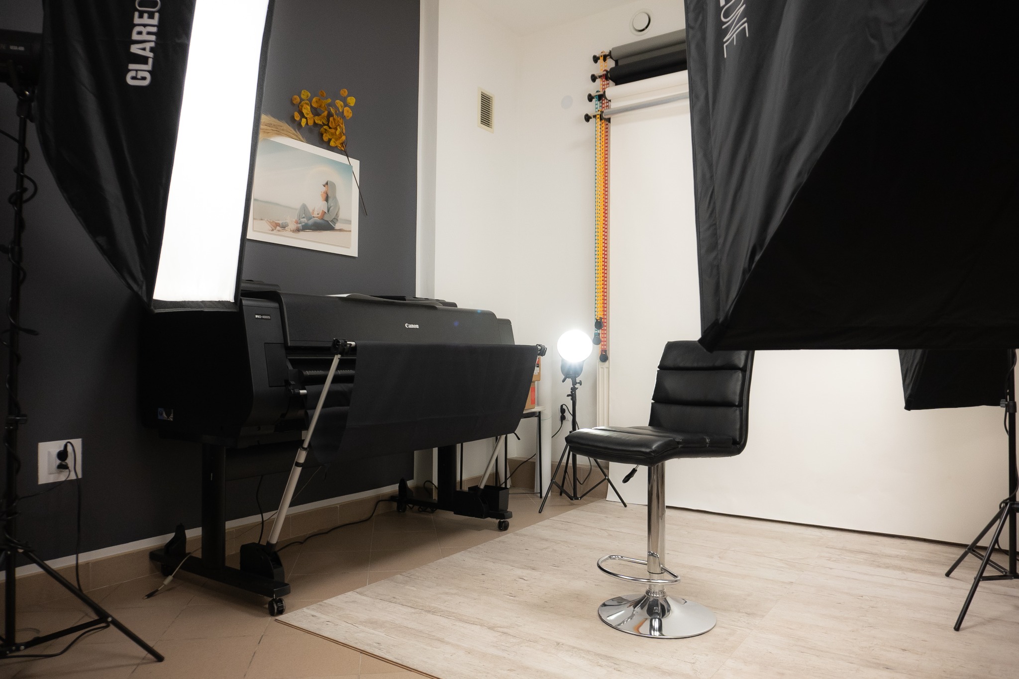 Studio fotograficzne z profesjonalnym sprzętem: lampy błyskowe z softboxami, ploter drukujący Canon, krzesło barowe i tła w rolkach.