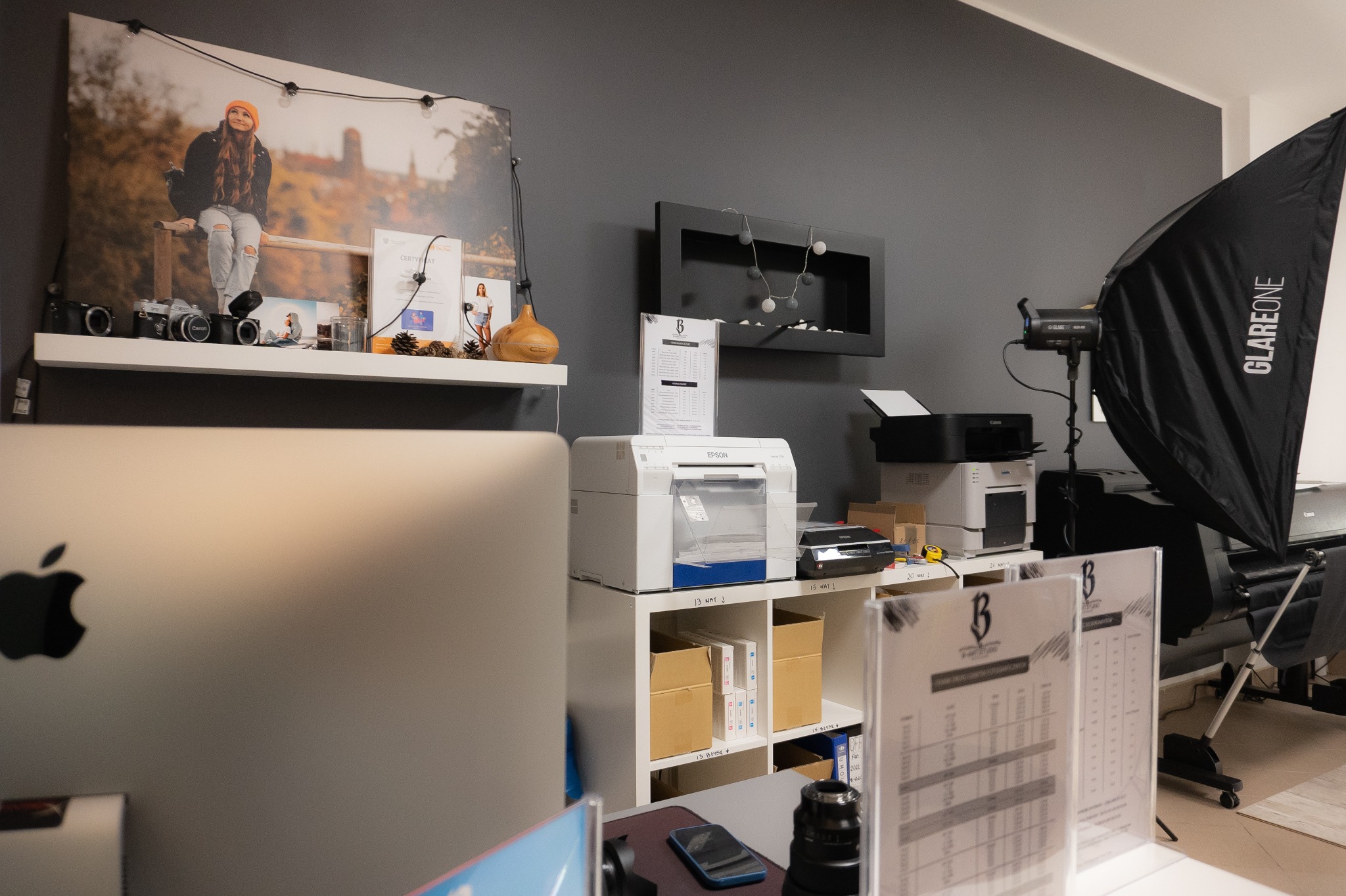Wnętrze studia fotograficznego: widoczny sprzęt oświetleniowy (softbox GlareOne), drukarki Epson i Canon, monitory Apple, półka z aparatami analogowymi i powiększone zdjęcie plenerowe kobiety...