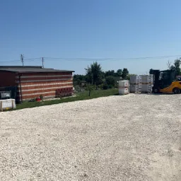 Żółty wózek widłowy na żwirowym placu z paletami białych, opakowanych grzejników, budynek w tle i błękitne niebo.