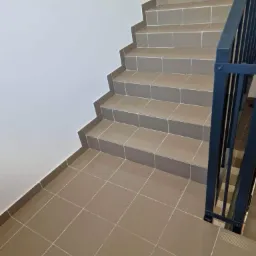 Wyłożone brązowymi płytkami schody z czarną metalową barierką, widok z góry ukazujący szczegóły fug i teksturę płytek.