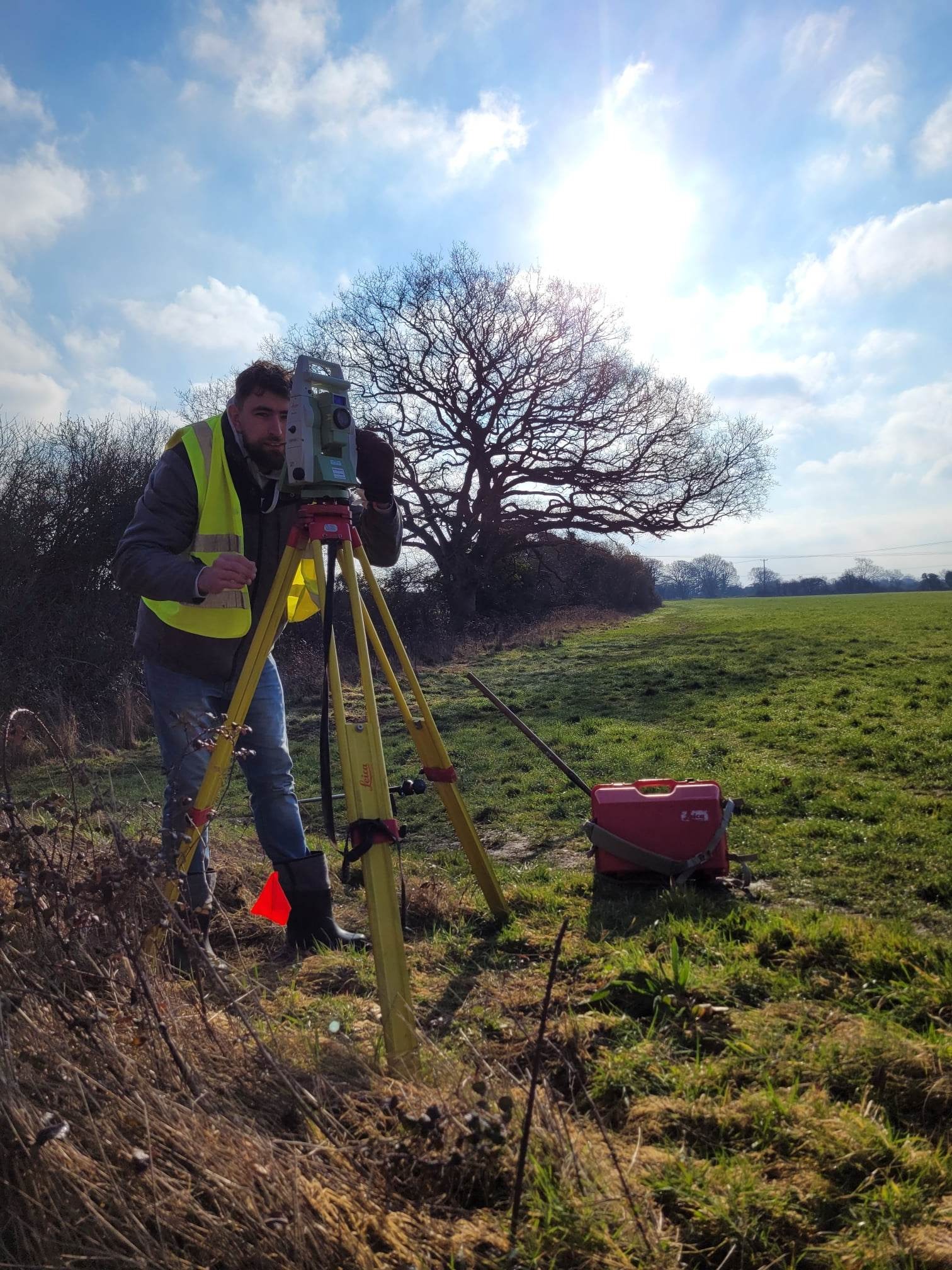 Mężczyzna w kamizelce odblaskowej obsługuje teodolit Leica na trójnogu, na tle zielonego pola i samotnego drzewa pod błękitnym niebem z chmurami.