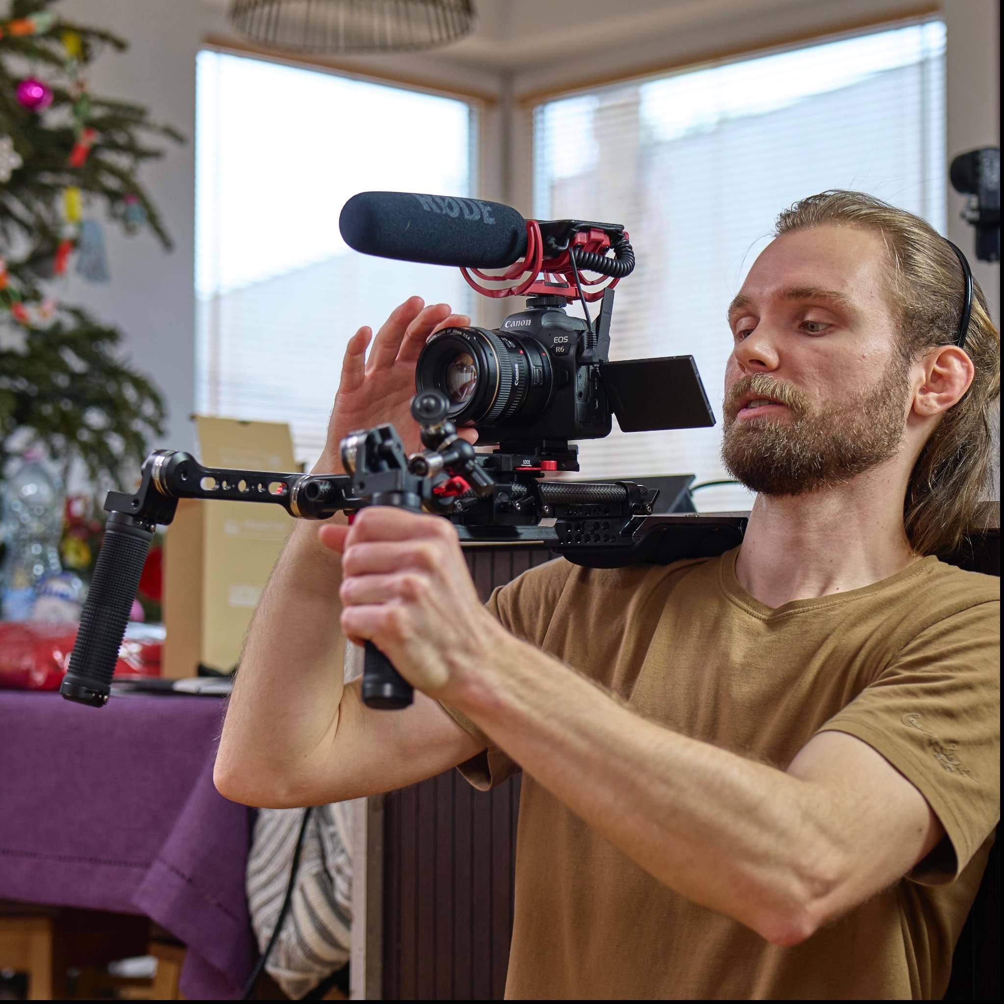 Mężczyzna z brodą obsługuje kamerę Canon EOS R6 z mikrofonem Rode na stabilizatorze, przygotowując się do nagrania w pomieszczeniu z choinką w tle.