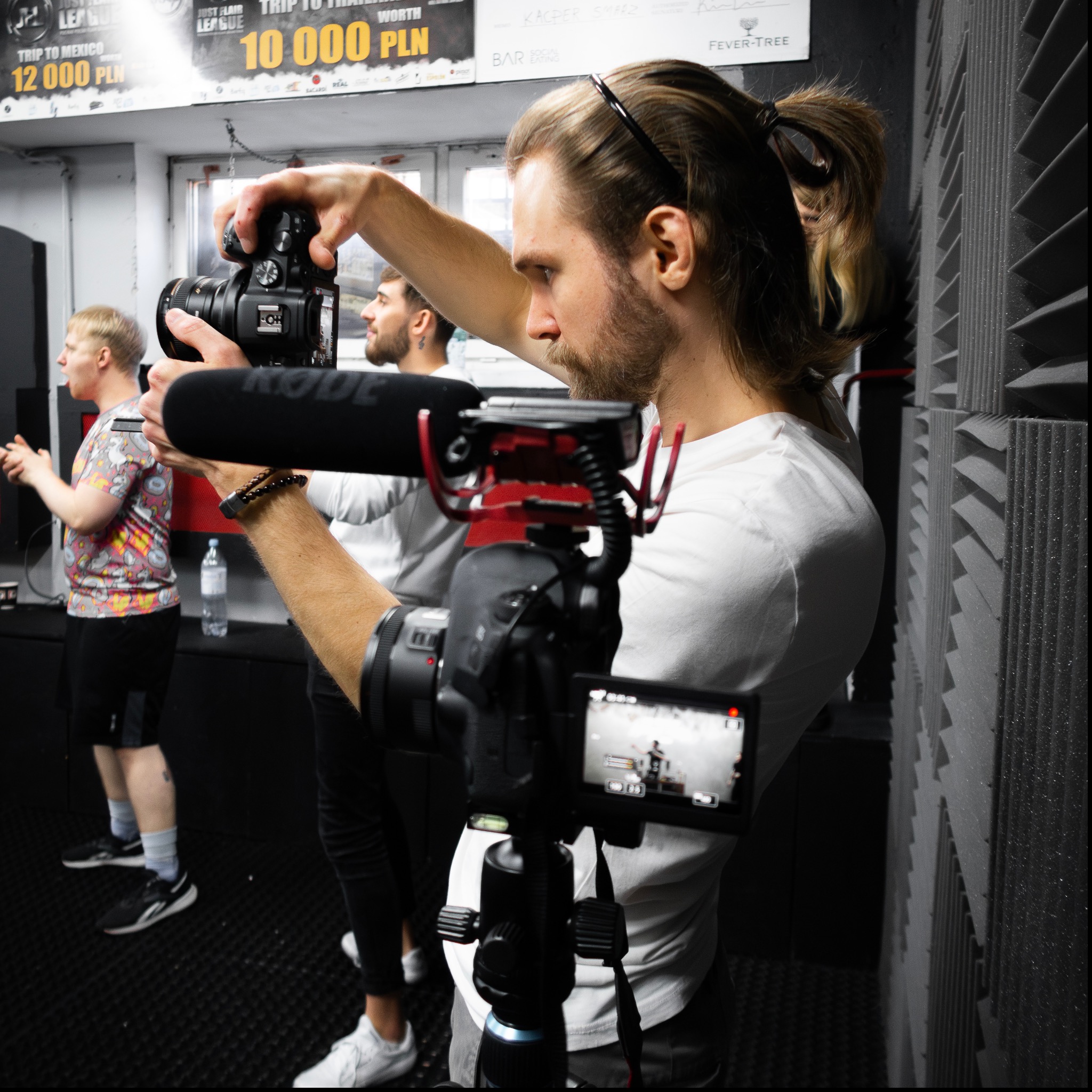 Ekipa filmowa podczas nagrywania w studio z panelami akustycznymi; widoczny operator kamery z mikrofonem Rode, kamera na statywie i aktorzy w tle. Na ścianie plakaty reklamowe.