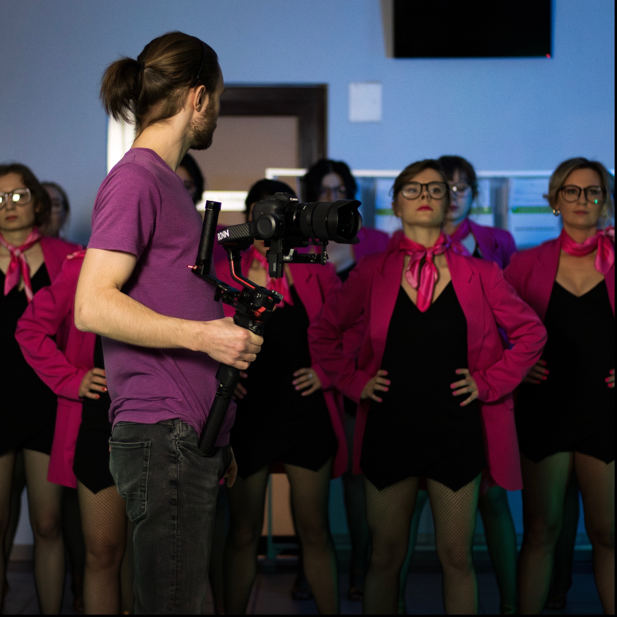 Operator z tyłu, trzymający kamerę na stabilizatorze, filmuje grupę kobiet w różowych marynarkach i czarnych sukienkach.