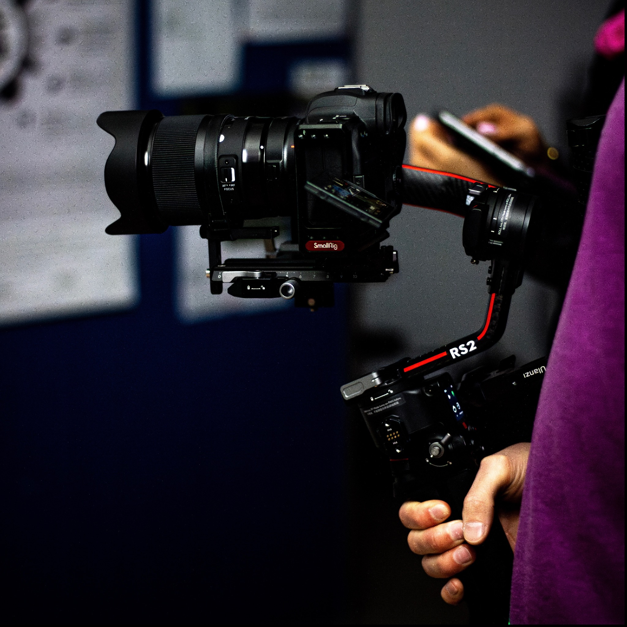 Profesjonalny aparat fotograficzny z obiektywem i stabilizatorem RS2 w dłoni operatora, w tle osoba korzystająca ze smartfona.