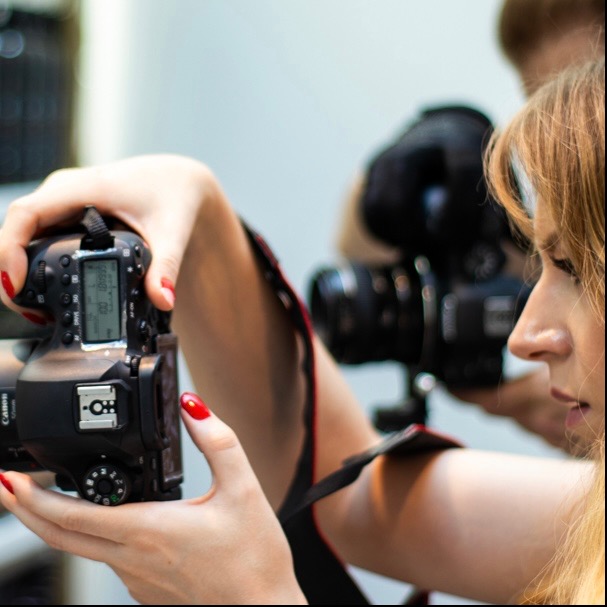 Kobieta z czerwonymi paznokciami trzyma aparat Canon DSLR, w tle drugi aparat na statywie, widok z profilu.