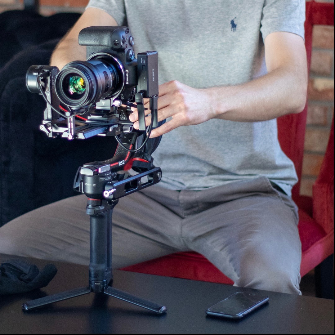Operator przygotowuje kamerę Canon EOS z obiektywem i stabilizatorem Ronin RS2 na statywie, smartfon leży na stole.