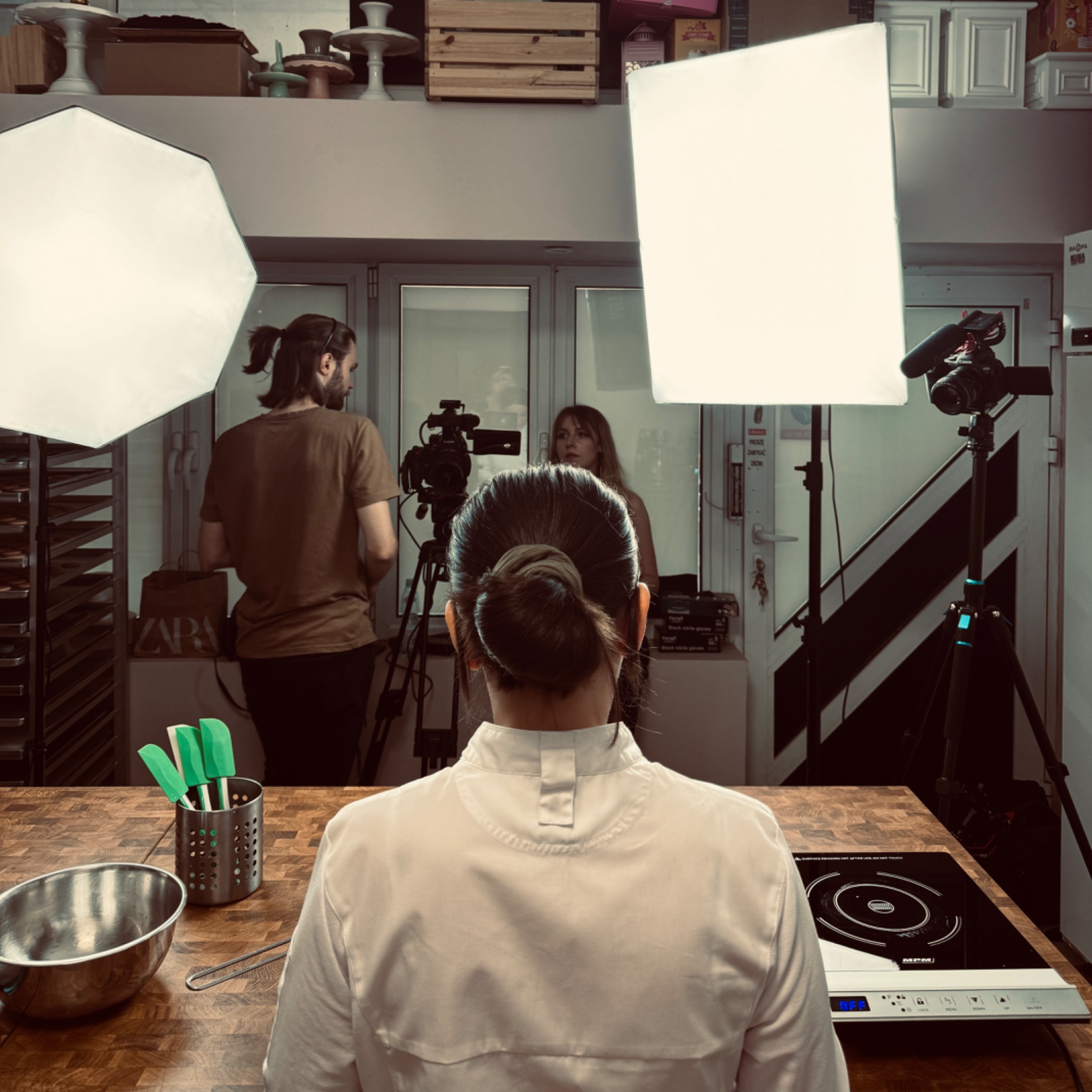 Ekipa filmowa podczas nagrywania sceny kulinarnej w studiu, oświetlenie studyjne, kamery na statywach, w tle półki z akcesoriami kuchennymi, na pierwszym planie stół z miską i przyborami kuchennymi.