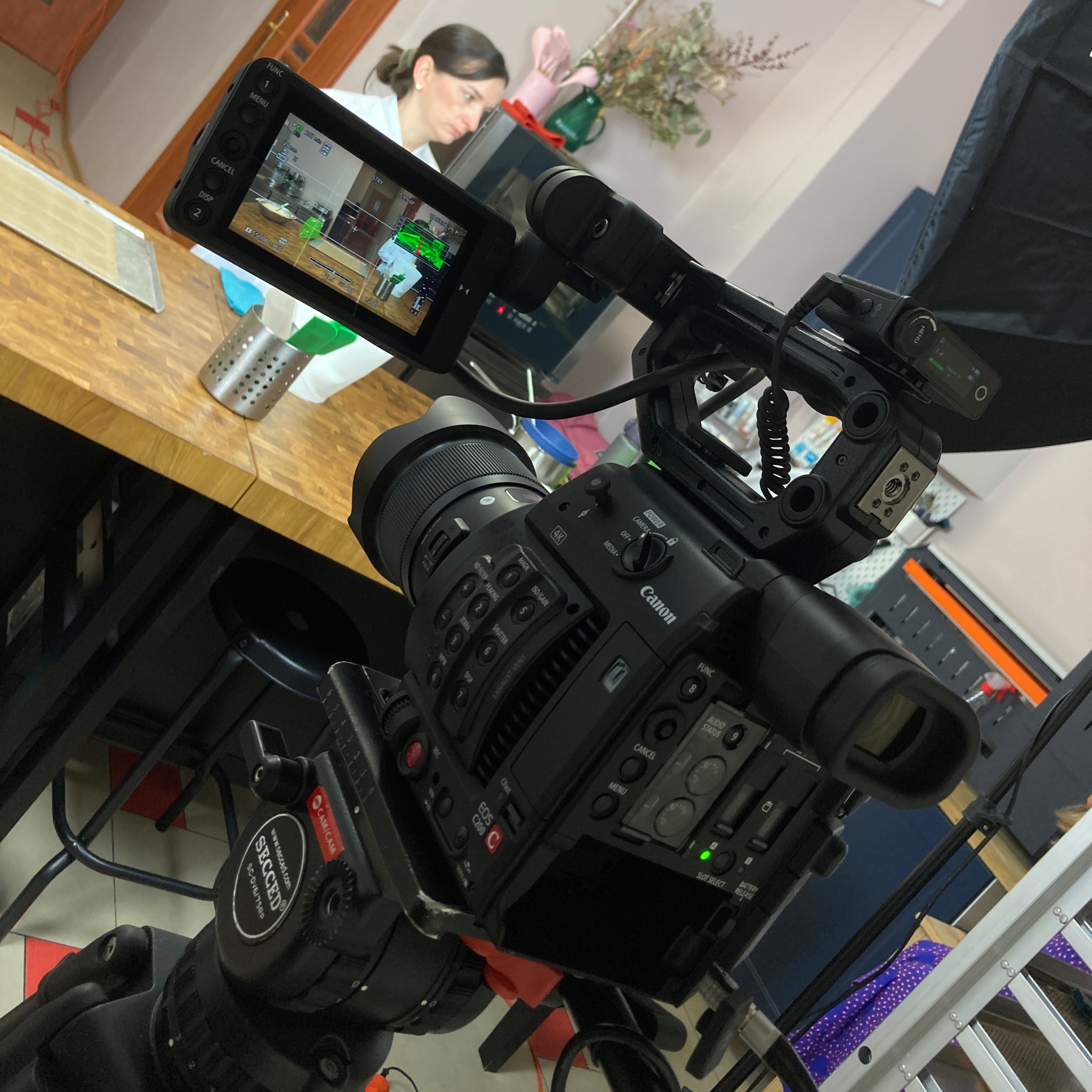 Profesjonalna kamera Canon EOS C200 na statywie Secced, skierowana na plan zdjęciowy z kobietą w białym fartuchu, widoczną na ekranie podglądu kamery, oraz akcesoriami kuchennymi na stole...