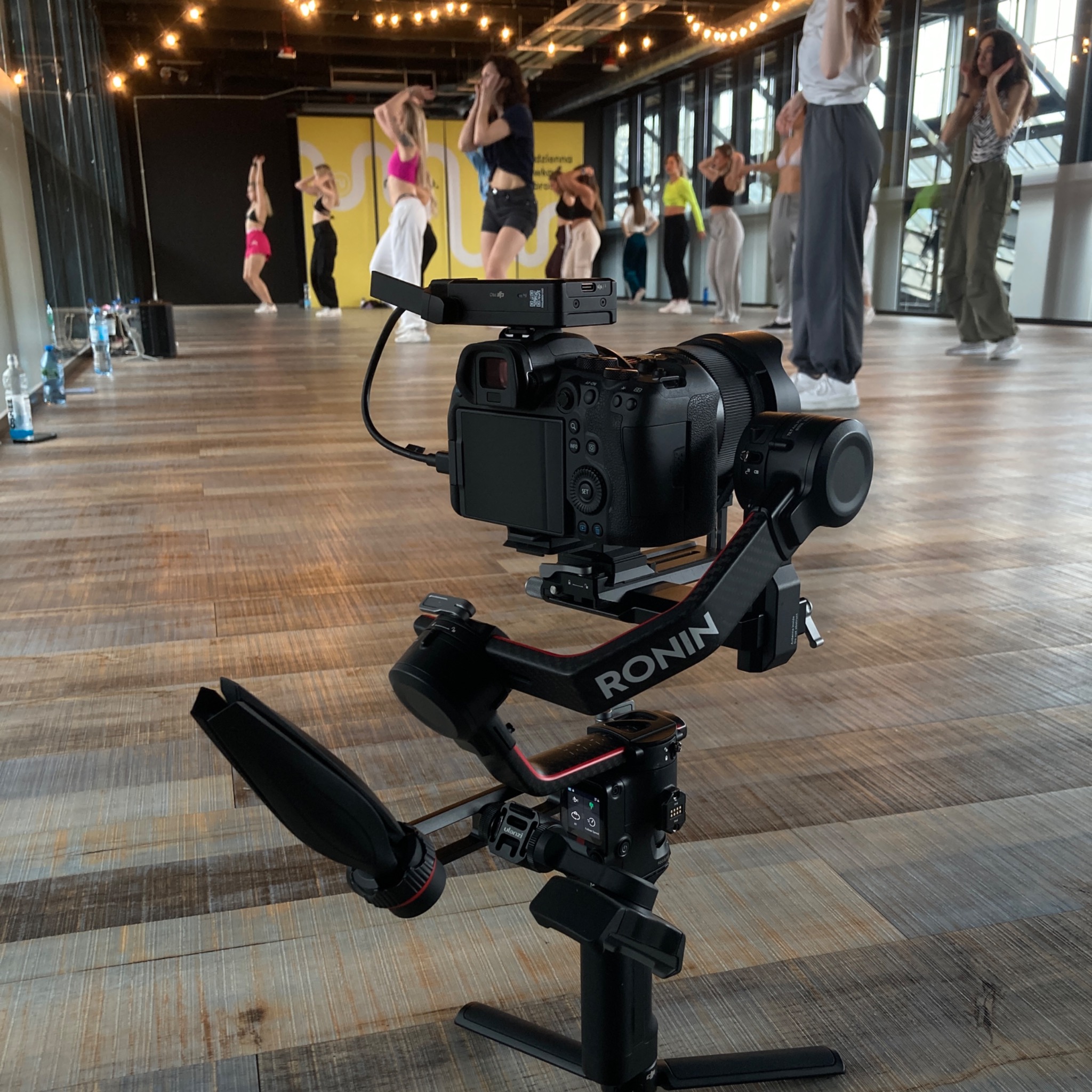 Kamera na stabilizatorze Ronin, skierowana w stronę grupy tancerzy w sali z drewnianą podłogą i oświetleniem girlandami.