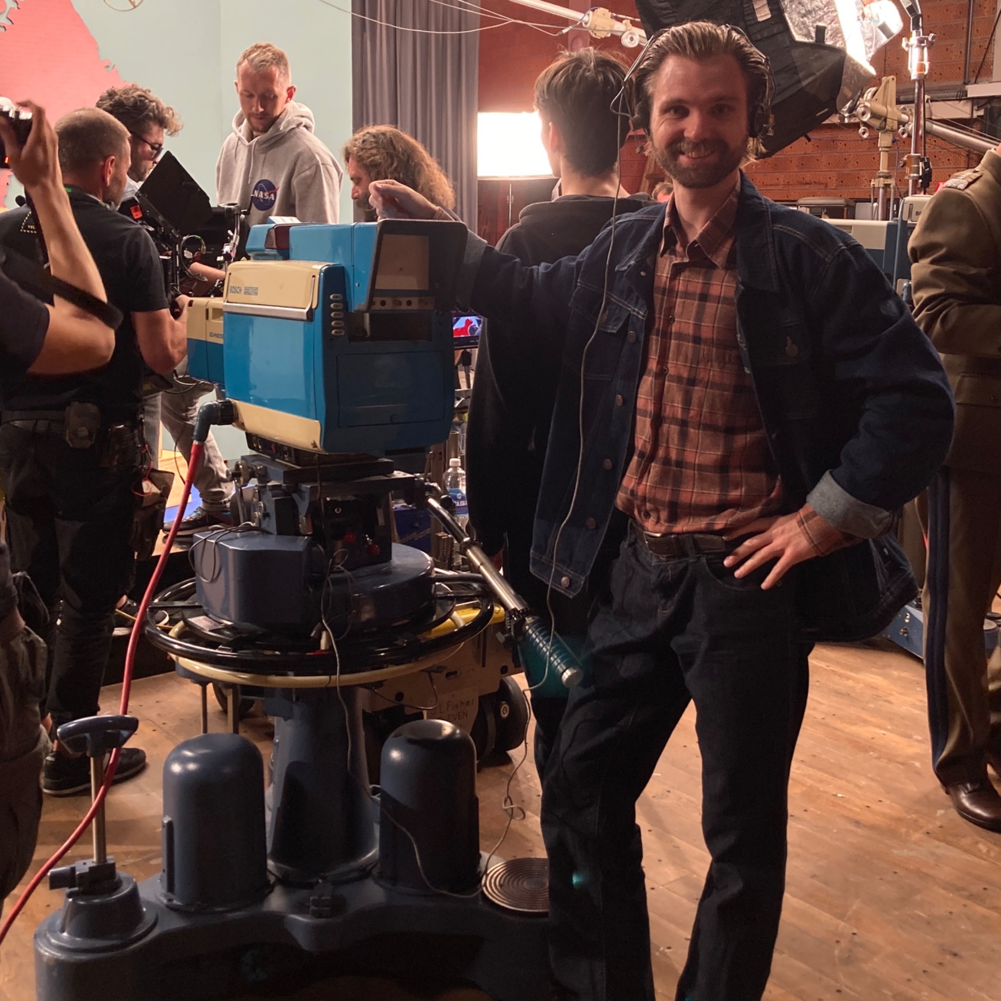 Mężczyzna w słuchawkach, w dżinsowej kurtce i kraciastej koszuli, opiera się o starą kamerę telewizyjną na planie filmowym, w tle ekipa filmowa i oświetlenie.