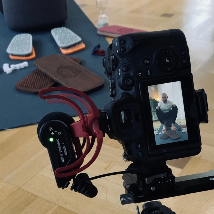 Kamera z mikrofonem nagrywa mężczyznę ćwiczącego na drewnianej desce do balansowania, akcesoria do ćwiczeń i szklana klepsydra w tle.