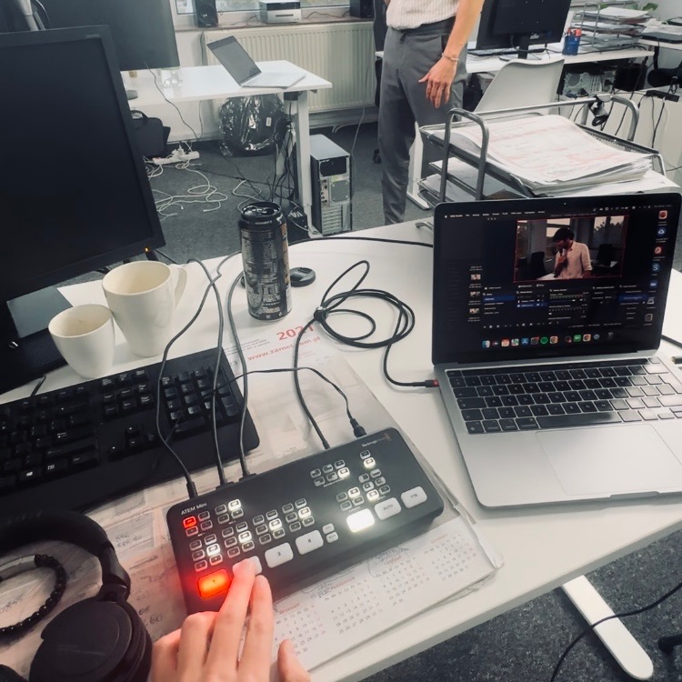 Stanowisko pracy z mikserem wideo ATEM Mini, laptopem z otwartym oknem programu do montażu, słuchawkami i kubkami na biurku w biurowym otoczeniu.