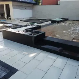 W trakcie układania jasnoszarej kostki brukowej o prostokątnym kształcie, obrzeża wykończone ciemną kostką, widoczne czarne donice i przygotowane podłoże w tle.