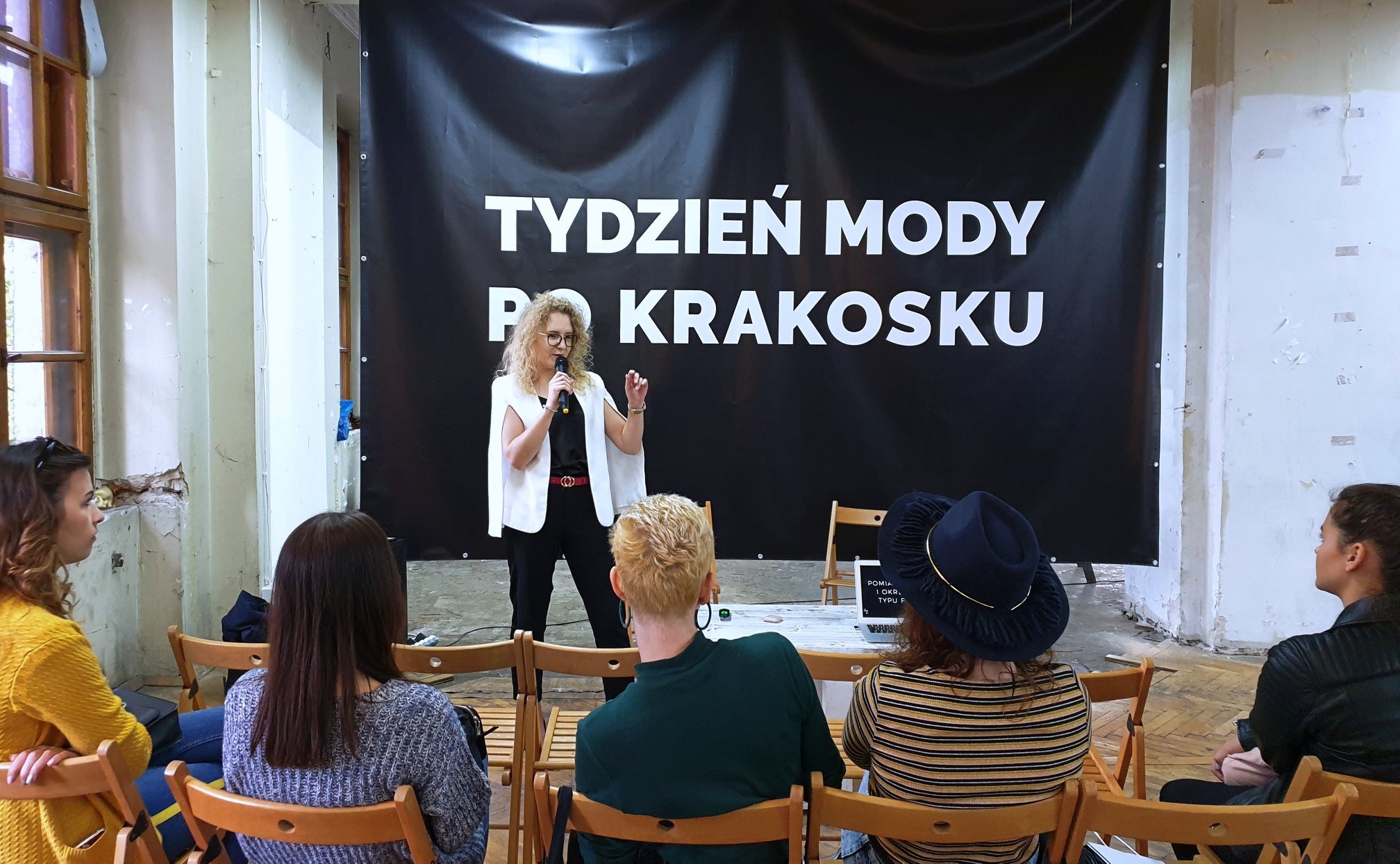 Prelegentka w białej marynarce i czarnych spodniach przemawia podczas Tygodnia Mody po Krakosku; widownia siedzi na drewnianych krzesłach.