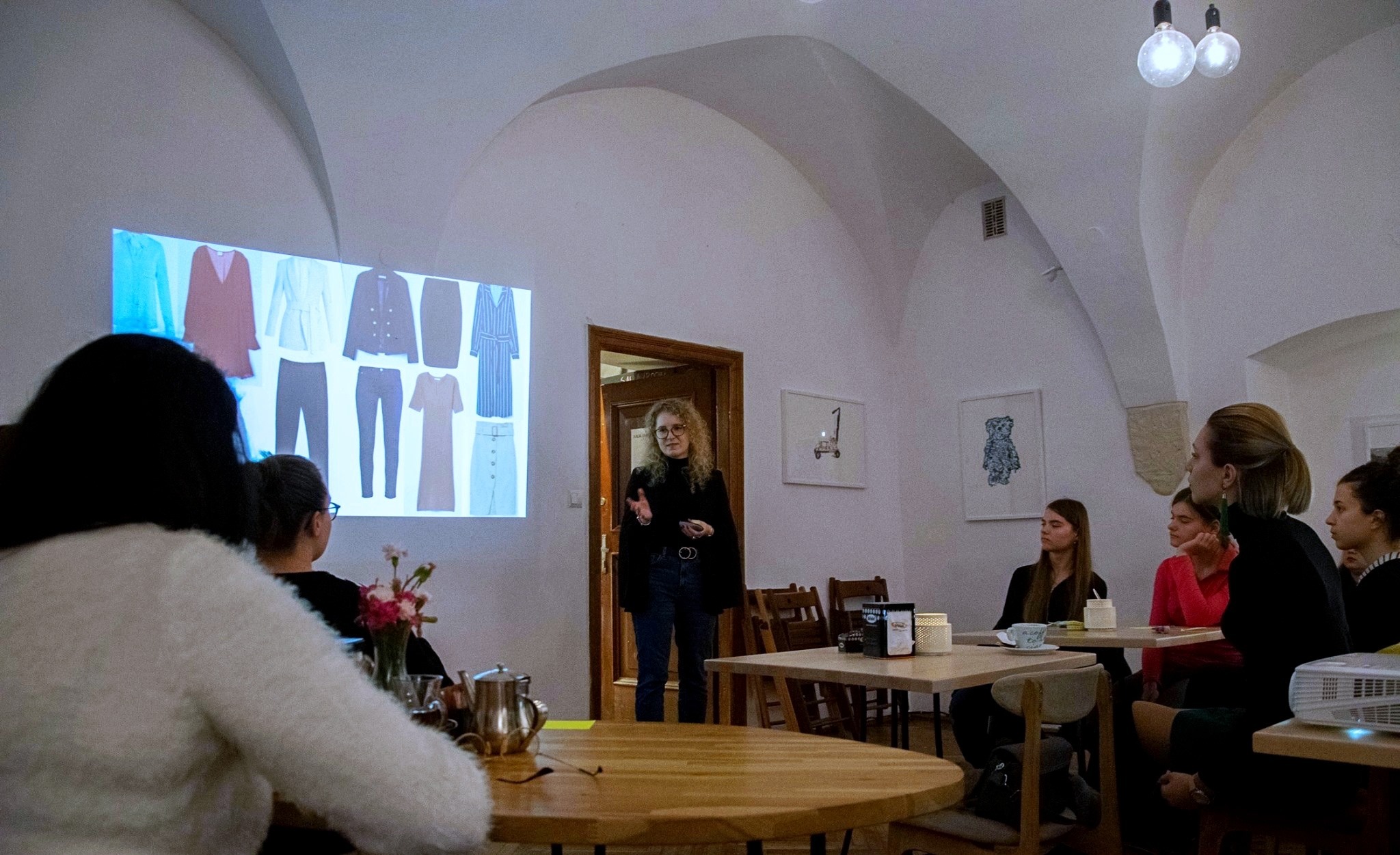 Prezentacja kolekcji ubrań na projektorze dla grupy osób w pomieszczeniu z łukowym sufitem i minimalistycznym wystrojem.