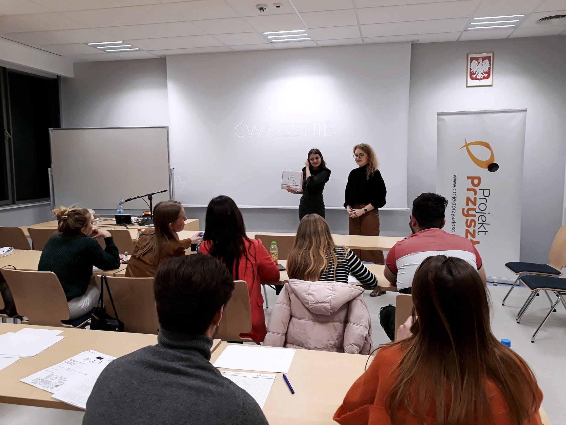 Sala szkoleniowa z uczestnikami siedzącymi przy stołach, dwie kobiety prowadzą ćwiczenie z użyciem tablicy z rysunkami, na ścianie napis 'Ćwiczenie' i flaga Polski.