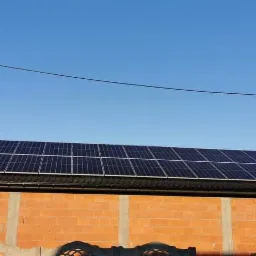 Panele słoneczne na dachu budynku z czerwonej cegły w słoneczny dzień. Widoczny fragment ogrodzenia.