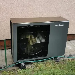 Szara pompa ciepła marki Nesheat, zainstalowana na kostce brukowej obok trawnika, z widocznym przewodem doprowadzającym.