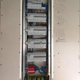 Otwarta skrzynka rozdzielcza z bezpiecznikami i przewodami w trakcie instalacji, widoczne oznaczenia pomieszczeń na ścianie obok.