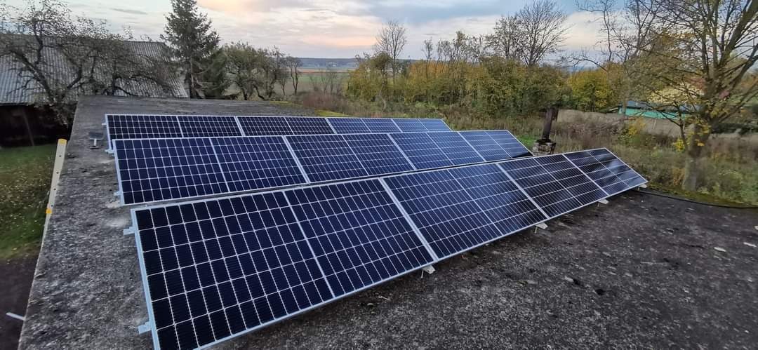 Rząd paneli słonecznych na płaskim, betonowym dachu budynku gospodarczego, z widokiem na jesienny krajobraz i zachmurzone niebo.