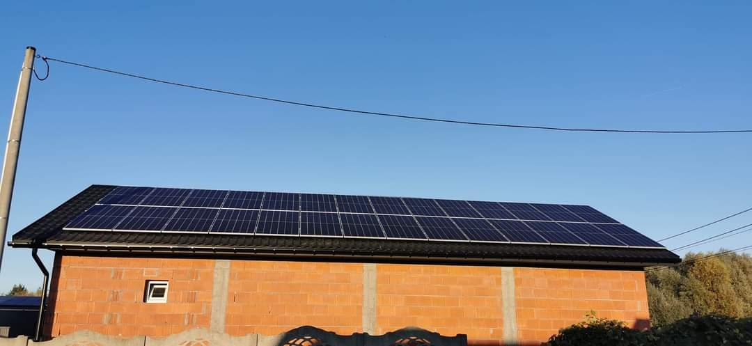 Panele słoneczne na dachu budynku z czerwonej cegły w słoneczny dzień. Widoczny fragment ogrodzenia.