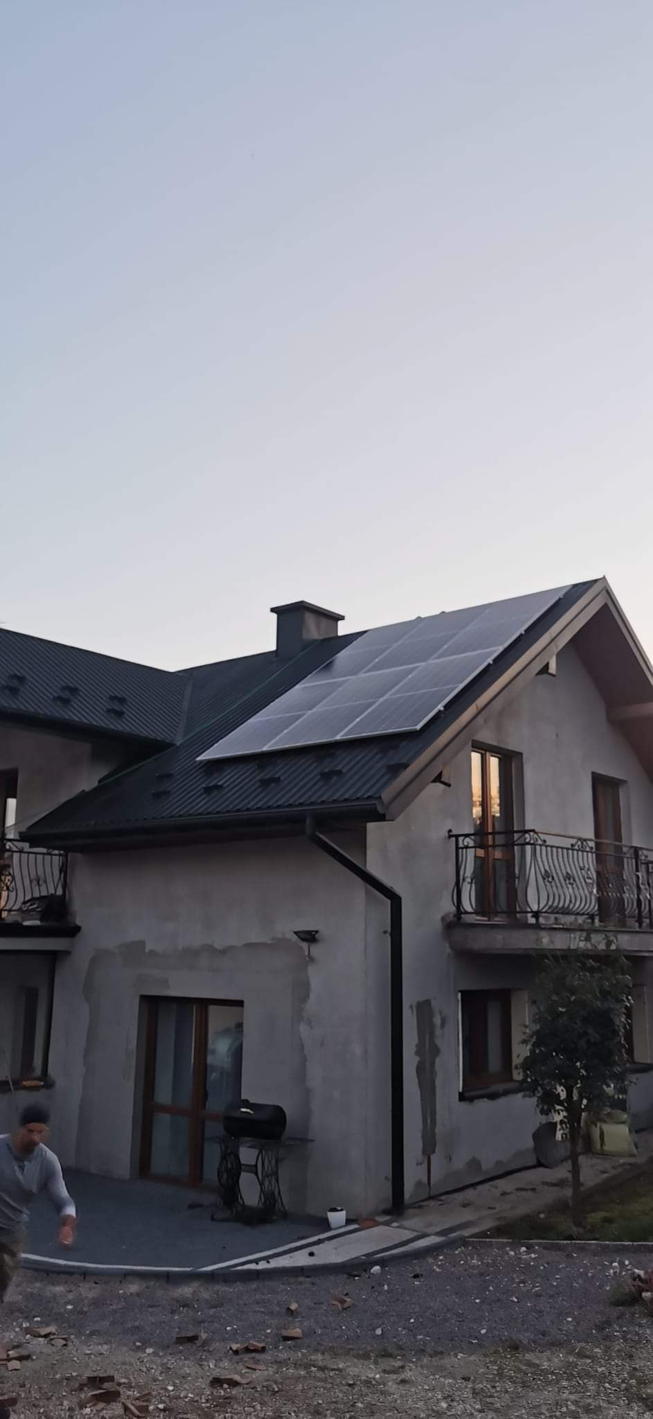 Dom jednorodzinny z panelami słonecznymi na dachu z czarnej blachy, widoczne okna z odbijającym się zachodem słońca, elewacja w trakcie wykończenia.