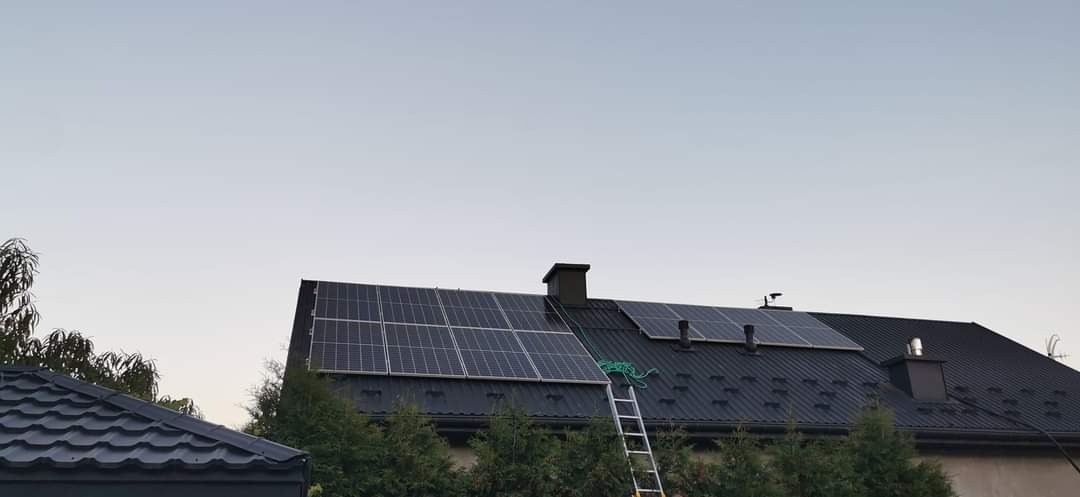 Panele słoneczne na dachu domu z blachy trapezowej, drabina oparta o dach, zielony wąż ogrodowy leżący na panelach.