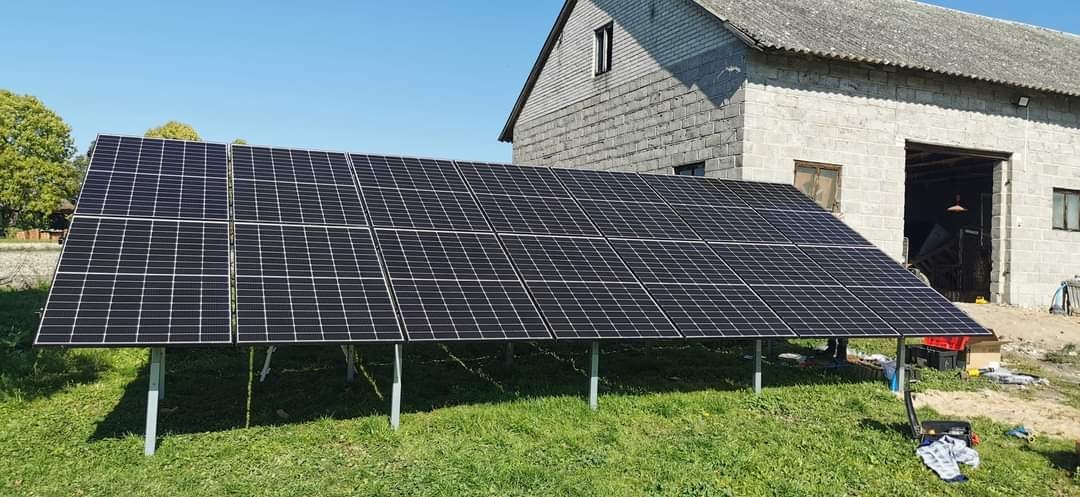 Instalacja paneli słonecznych na konstrukcji wsporczej na trawie, w tle budynek gospodarczy z bramą, narzędzia i skrzynki obok.