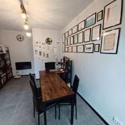 Wnętrze biura z drewnianym stołem, czarnymi krzesłami, ścianą udekorowaną certyfikatami i dyplomami w ramkach, widoczny robot sprzątający i wiszący na ścianie panel grzewczy.