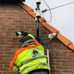 Instalacja zewnętrznych kamer monitoringu na ścianie budynku z cegły, widoczny pracownik w odblaskowej kurtce z logo firmy na drabinie, słup z izolatorami kabli.