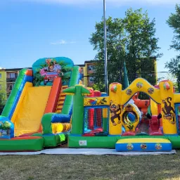 Kolorowy, dmuchany plac zabaw z motywem dżungli, składający się ze zjeżdżalni i zamku, ustawiony na trawie przed budynkiem mieszkalnym i ogrodzeniem.