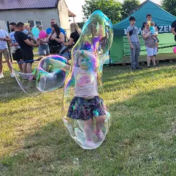 Dziewczynka uśmiecha się, stojąc wewnątrz ogromnej, mieniącej się bańki mydlanej na trawie, w tle widoczni ludzie i zielony namiot.