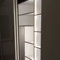 Wnętrze szafy wnękowej z przesuwnymi drzwiami, oświetlone pionowymi paskami LED, z półkami o różnych wysokościach.