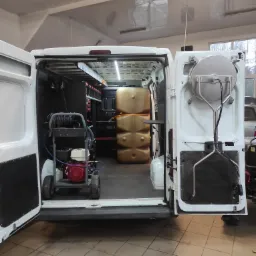 Wnętrze białego furgonu z otwartymi drzwiami, wyposażonego w zbiorniki na wodę, zwijak na wąż ciśnieniowy oraz myjkę ciśnieniową z silnikiem spalinowym.