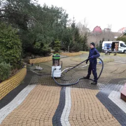 Pracownik w niebieskim kombinezonie czyści kostkę brukową wzorem w paski przy użyciu profesjonalnego odkurzacza wodnego, widoczny firmowy bus w tle.