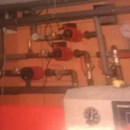 Kotłownia z widocznymi pompami obiegowymi, rurami, zaworami i manometrami na tle pomarańczowej ściany.